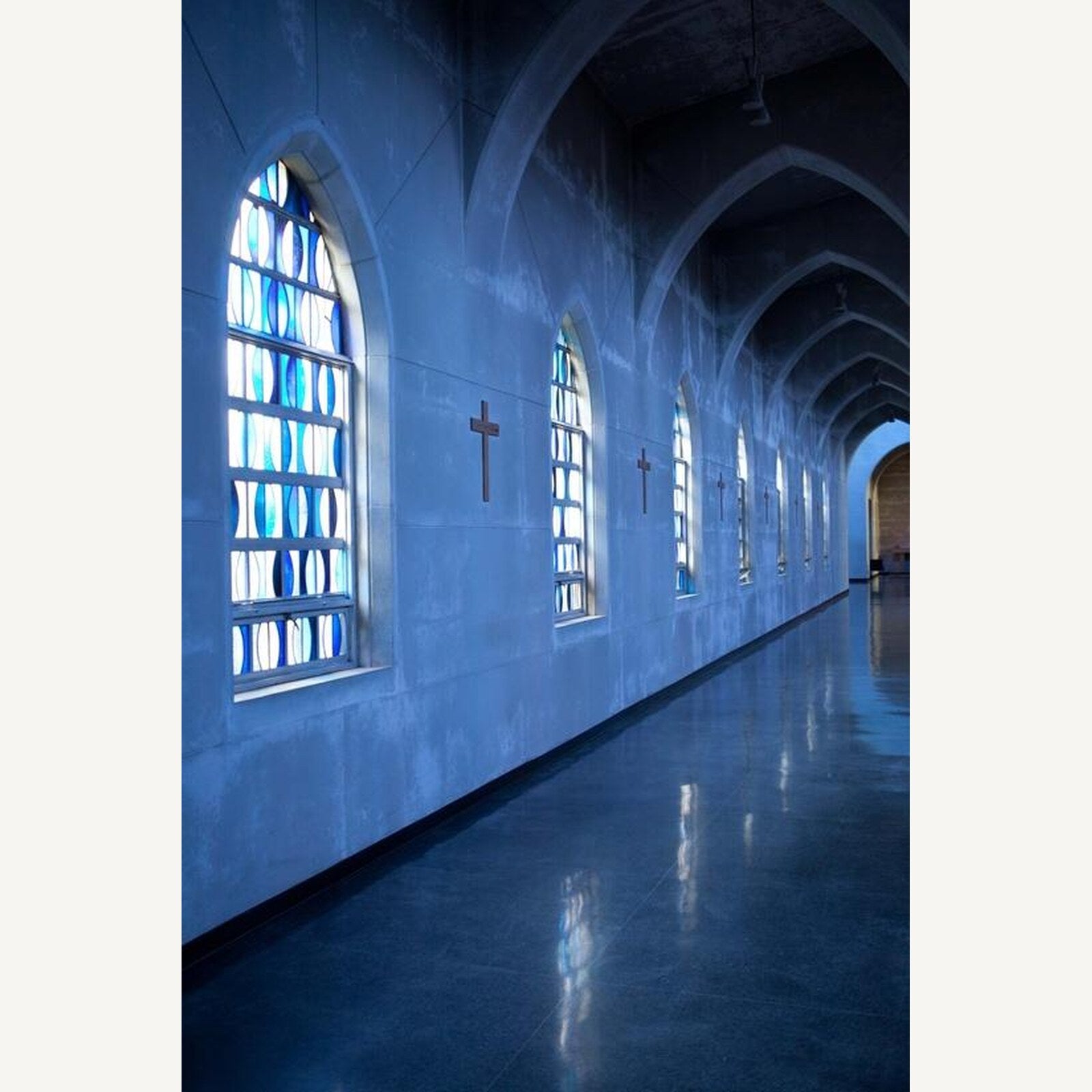 The Abbey Church cloister walk, a colonnade of pointed concrete arches with blue leaded-glass lancets and plain crosses along the wall.